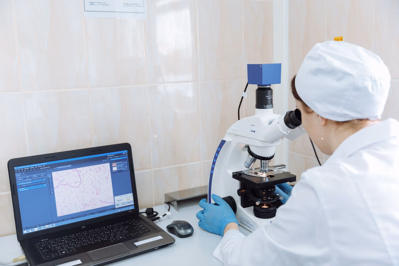 A laboratory technician working at a microscope beside a laptop